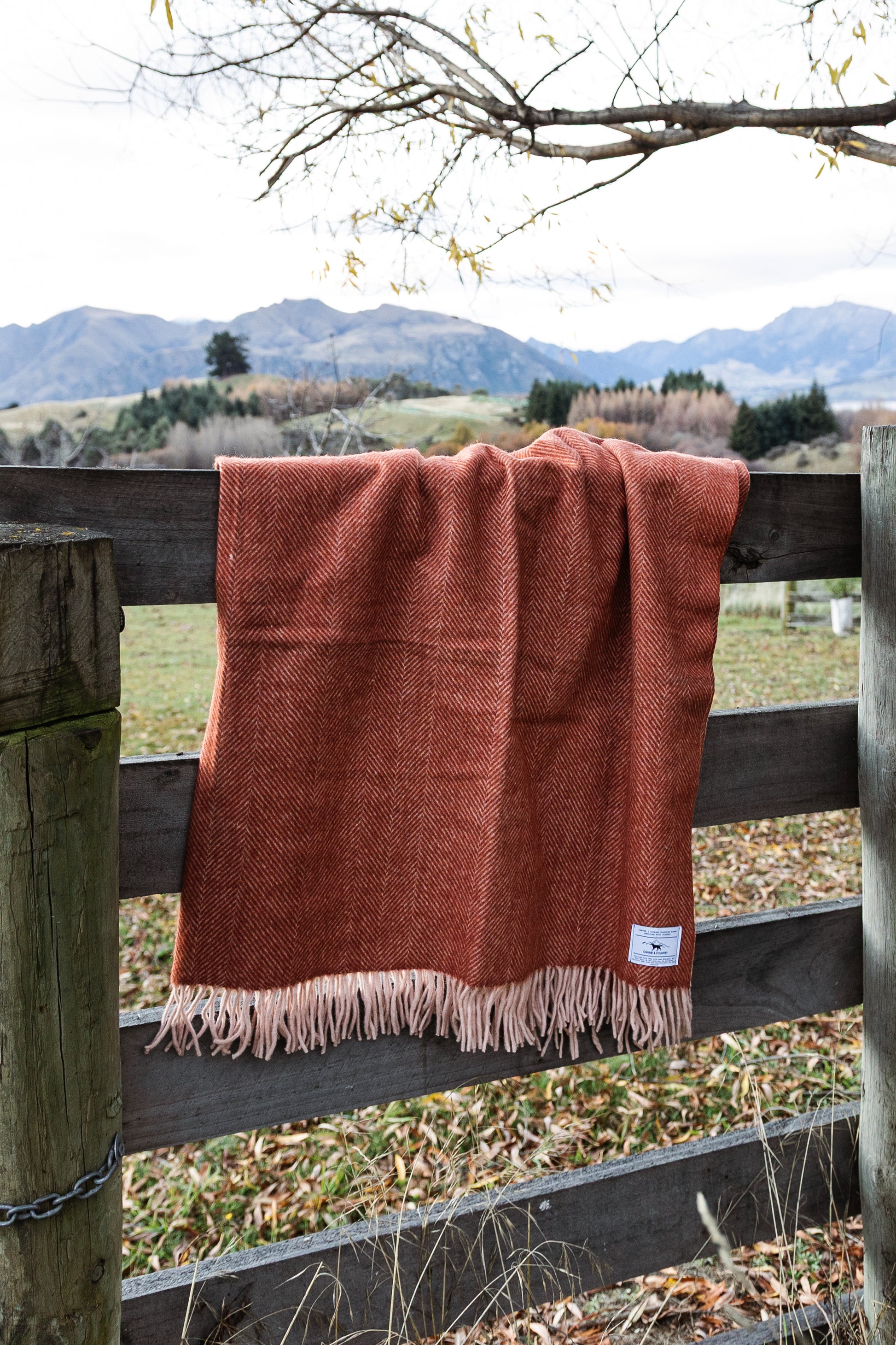 Rust coloured recycled wool blanket draped over a wooden fence, with a scenic country setting.