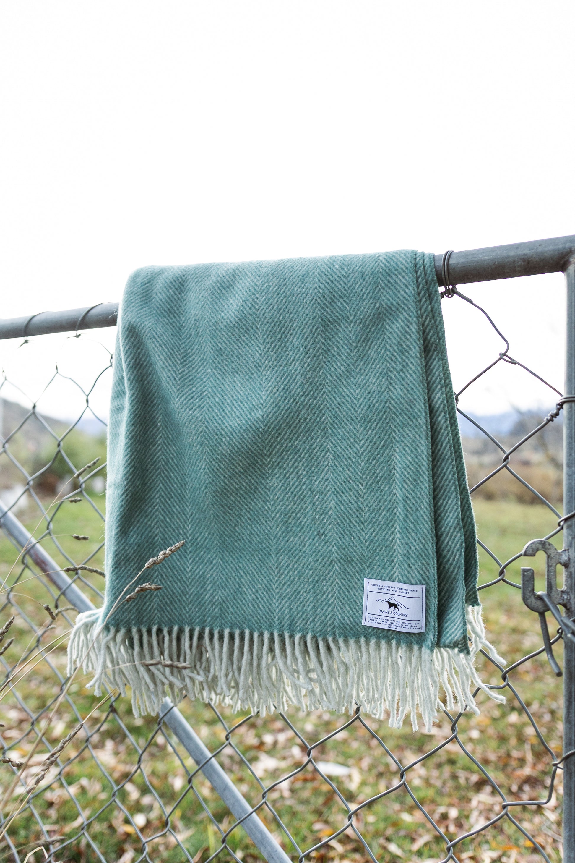 Recycled wool blanket with fringes draped over a chain-link fence in an country setting.