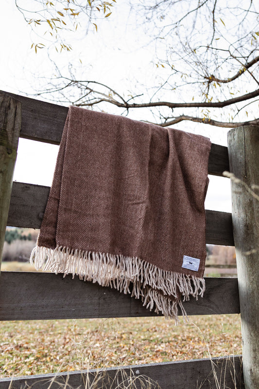 Brown herringbone blanket with fringes draped over a wooden fence outdoors